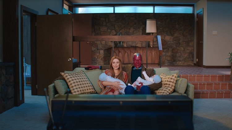 Elizabeth Olsen and Paul Bettany in a domestic scene from WandaVision, with both characters sitting on the sofa in front of the tv, each holding one of their baby twins.