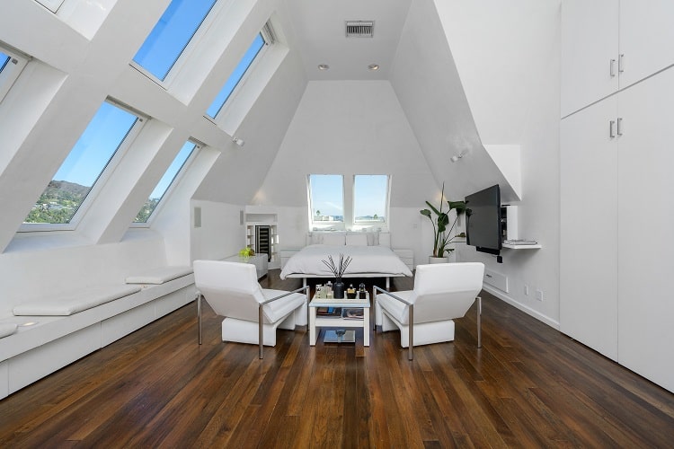 bedroom with a bed in the middle, sitting under vaulted ceilings with two rows of windows, with two white chairs