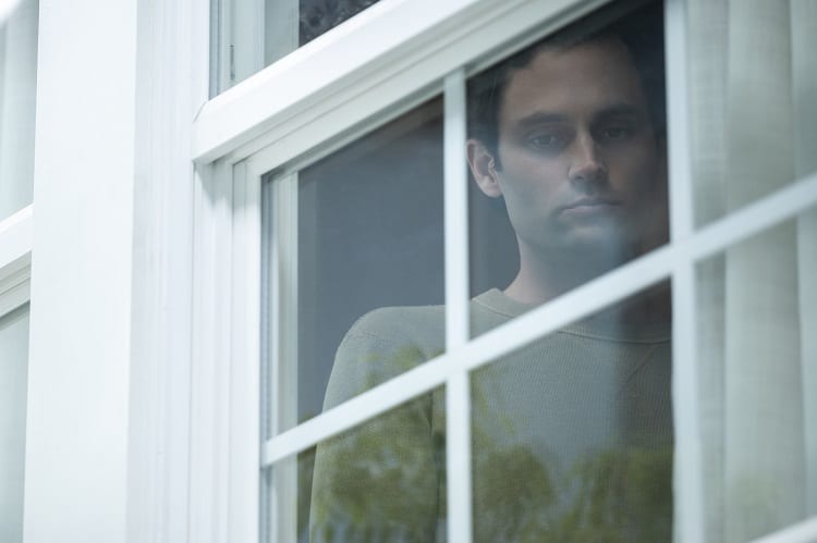 Penn Badgley as Joe Goldberg standing in the window of the house from Season 3 of You.