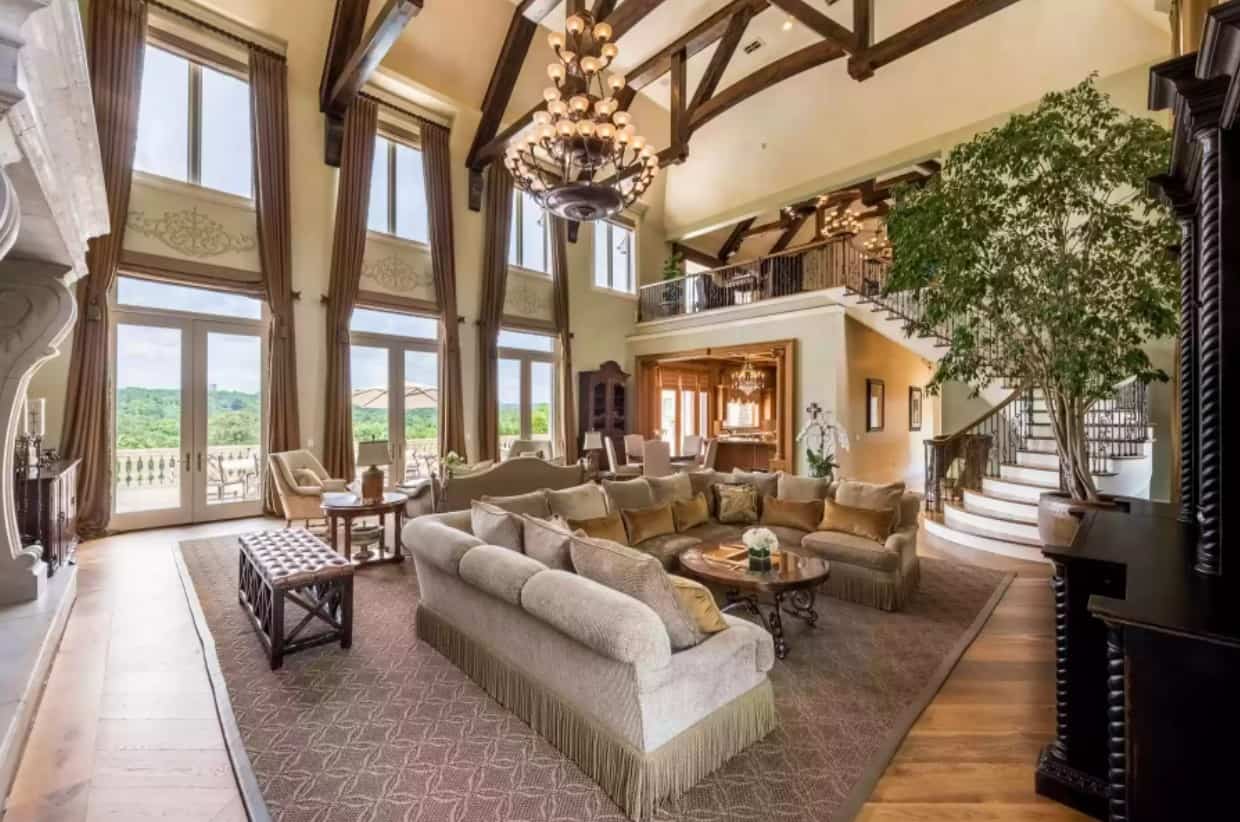 The grand, two-story living room has beamed wood ceilings, an opulent chandelier, and double-story windows.