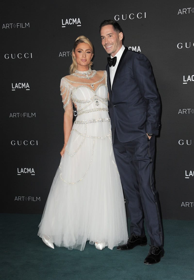 Paris Hilton and husband Carter Reum in formal attire, her wearing a heavily bedazzled white dress, him a dark navy suit