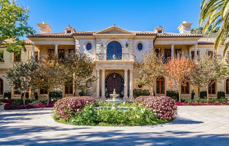 The entrance to Paris Hilton's house, with a circular fountain in front with many colorful plants and flowers