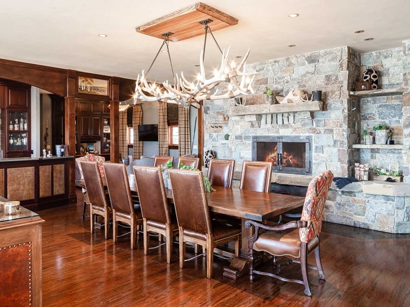 elegant-rustic dining area with a massive wood dining area, tufted leather chairs, a rock fireplace and antler chandelier