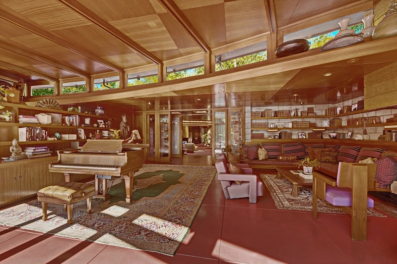 inside Frank Lloyd Wright's Tirranna House, where a warm wood-clad living room has a piano, sofa, chairs and lots of shelves