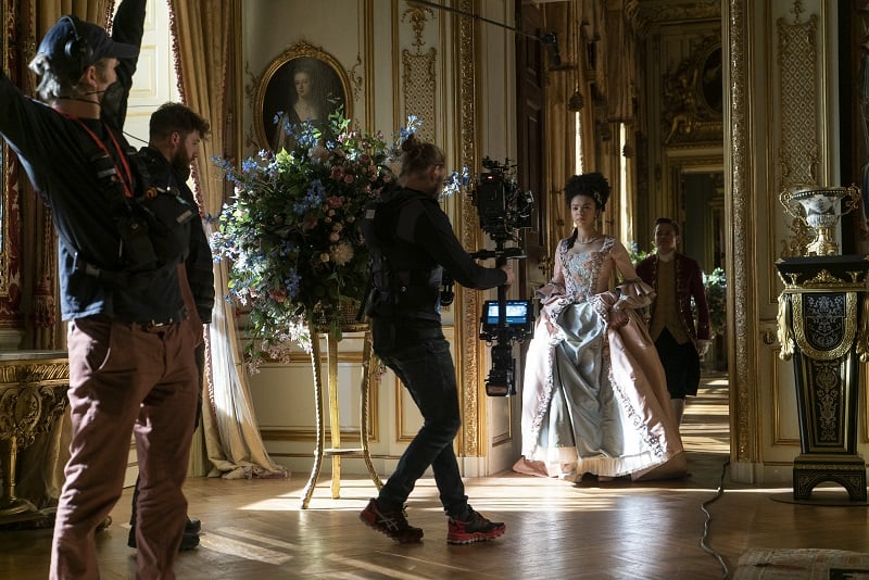 India Amarteifio as Young Queen Charlotte, Sam Clemmett as Young Brimsley in Queen Charlotte: A Bridgerton Story walking in the golden hallways of the castles, with the filming crew in front of them