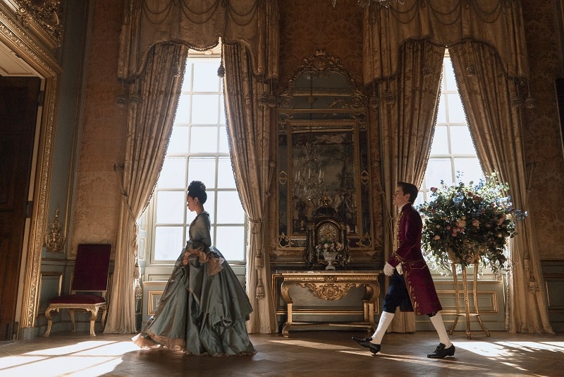 India Amarteifio as Young Queen Charlotte, Sam Clemmett as Young Brimsley in episode 102 of Queen Charlotte: A Bridgerton Story, walking through an opulent hallway inside the Buckingham House