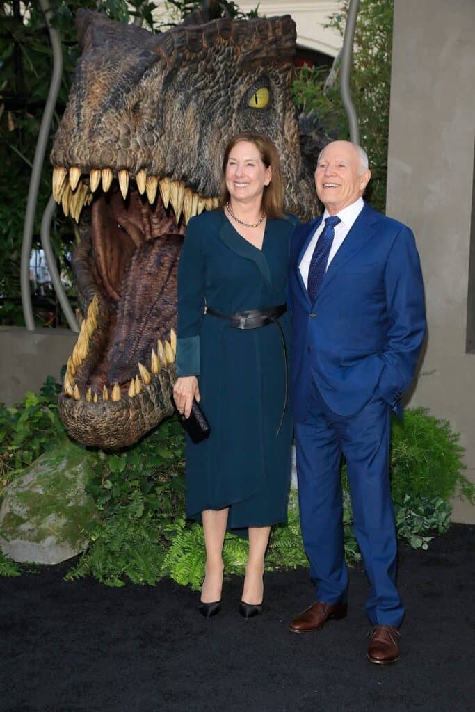 Kathleen Kennedy, Frank Marshall at the World Premiere of Jurassic World Dominion in 2022, next to a dinosaur replica with open mouth and sharp teeth. 