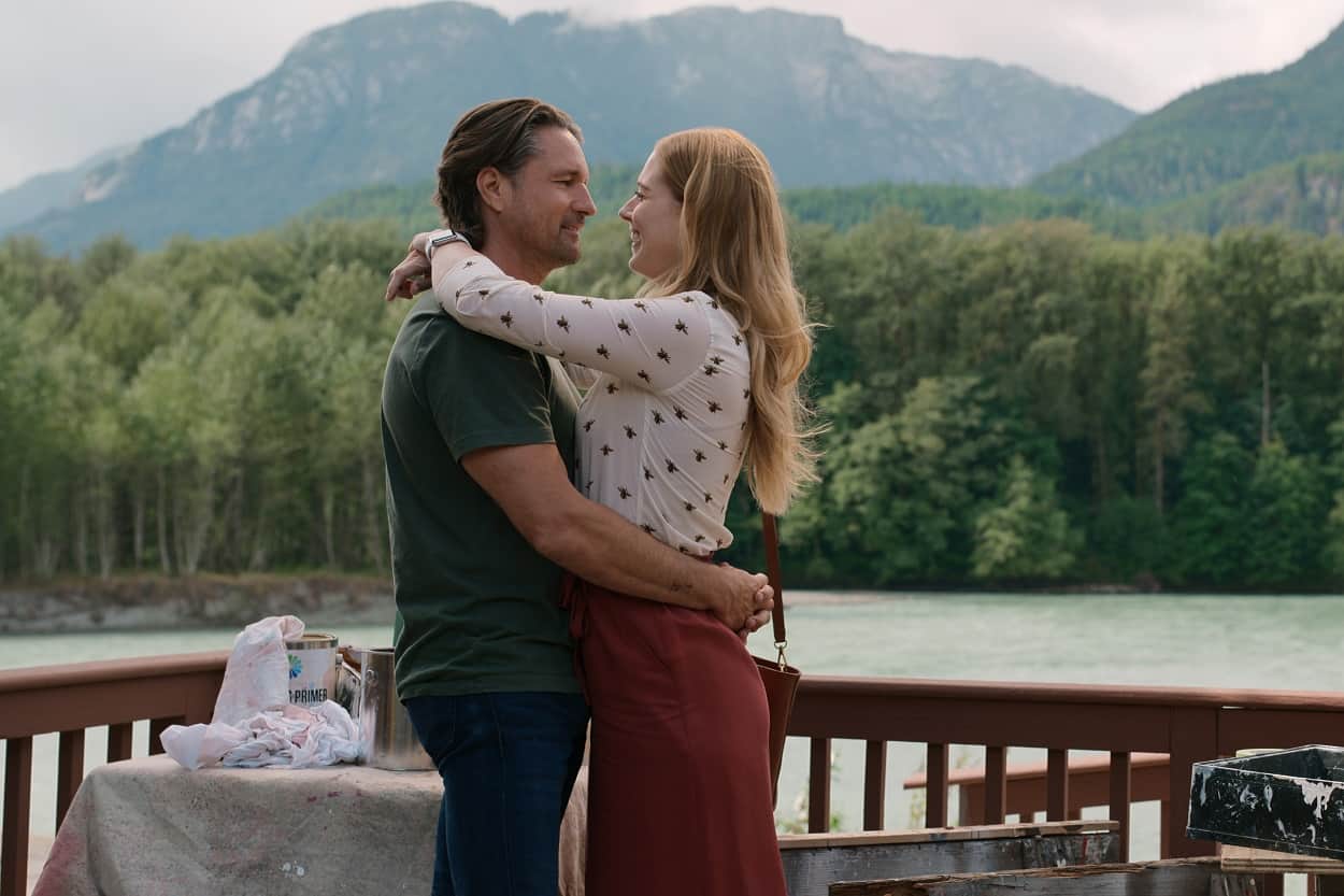 Virgin River scene showing Martin Henderson as Jack Sheridan, Alexandra Breckenridge as Mel Monroe in episode 404 of Virgin River. Scene takes place on the deck of Mel's cabin, with the lake and mountains in the background