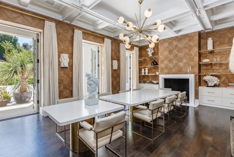 The formal dining room has coffered ceilings, brown patterned walls, a white table with chairs, and an elegant fireplace.