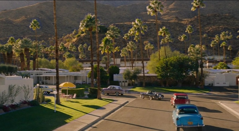 Scene showing the Victory neighborhood in Don't Worry Darling, surrounded by palm trees and with colorful vintage cars driving away.