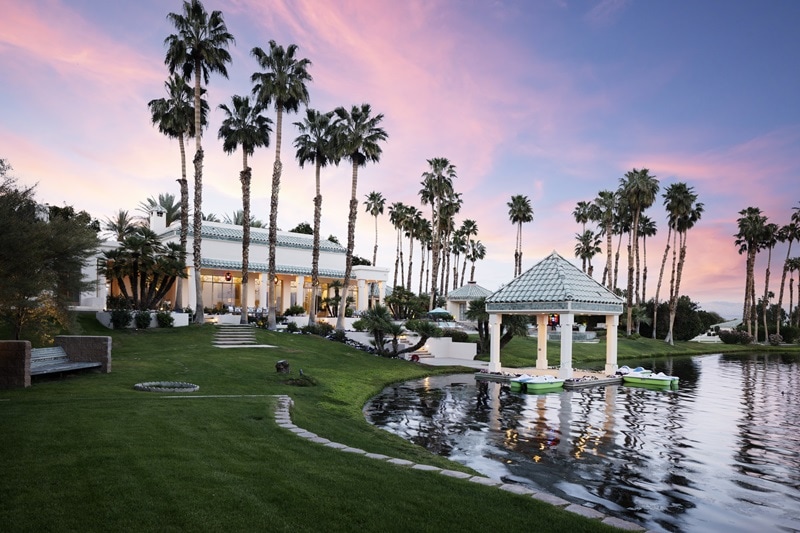 the main house on the property, a Moroccan-inspired luxury house seen from the backyard with lush lawns, lots of palm trees, and a man-made lake