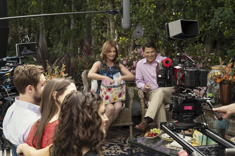 A behind-the-scenes shot showing the main characters in the TV series The Baxters gathered around in the backyard, opening presents with the filming cameras in view.