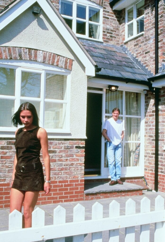 David Beckham standing at the entrance of the house, watching Victoria Beckham leaving, with a white picked fence in front.