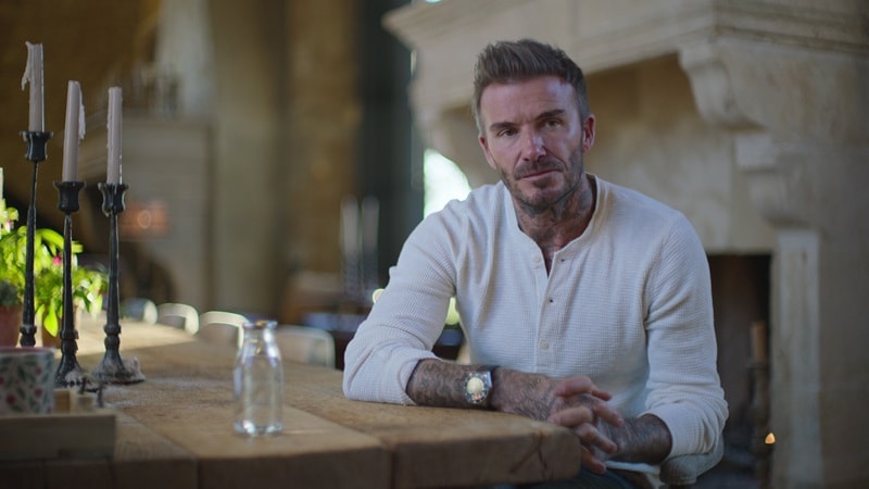 David Beckham standing at a table in his dining room with a stone fireplace behind him