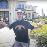 YouTube creator FaZe Rug standing in front of his California mansion, with arms extended, inviting viewers in.