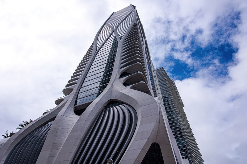 a closer view of the curving exoskeleton of the One Thousand Museum building in Miami, seen against the backdrop of a cloudy sky.