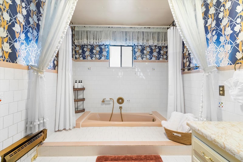 1980s-themed bathroom with a pink/orange sunken bathtub, floral wallpaper and white curtains. 