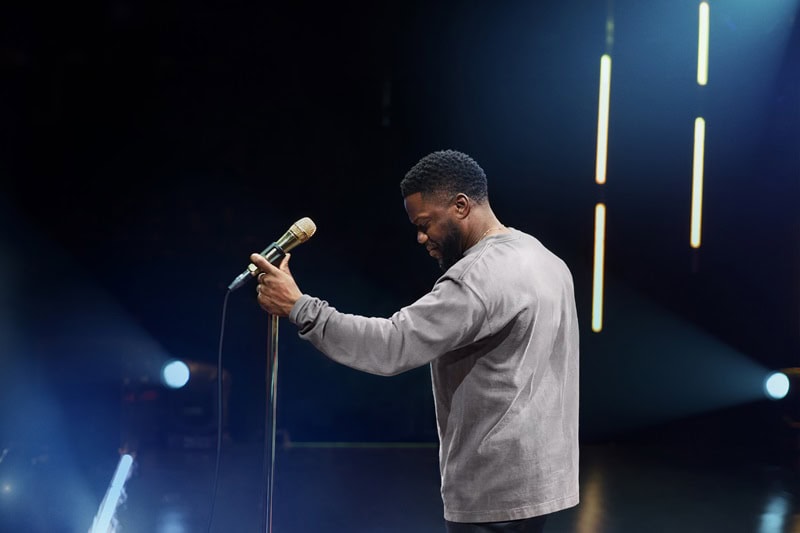 Actor/comedian Kevin Hart standing on a lit-up stage, holding his hands on a microphone and looking down while wearing a grey sweatshirt.