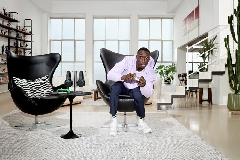 TikTok sensation Khaby Lame sitting on a revolving chair, gaming controller in hand, playing video games in a modern loft apartment with floor-to-ceiling windows. 