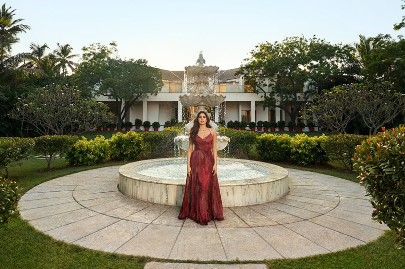 Janhvi Kapoor's house in Chennai, India. Janhvi is sitting in front of a water fountain, wearing a dark red dress, with the house visible in the background, surrounded by trees and shrubbery. 