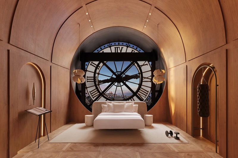 a bed set up in Musée d’Orsay's clock room, with a queen sized med in the middle, surrounded by wood paneling all around, with the clock in the background and Paris scenes visible through it. 