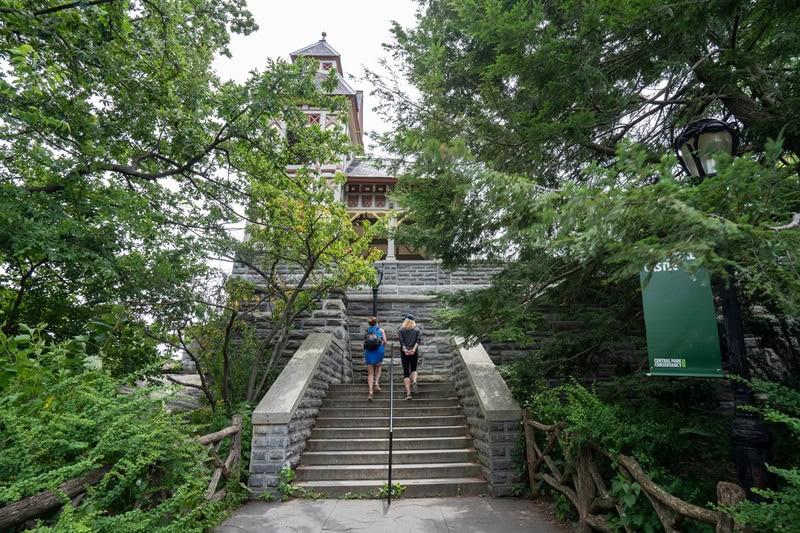 two women leisurely climbing the steps to the Belvedere Castle, while glancing up and admiring the architecture. The steps and the castle are surrounded by lush greenery and trees.