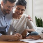 A couple smiling while signing a home purchase agreement, with a female lawyer's hand hovering over the document.