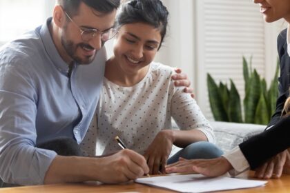 A couple smiling while signing a home purchase agreement, with a female lawyer's hand hovering over the document.