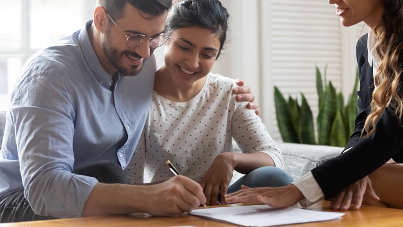 A couple smiling while signing a home purchase agreement, with a female lawyer's hand hovering over the document.