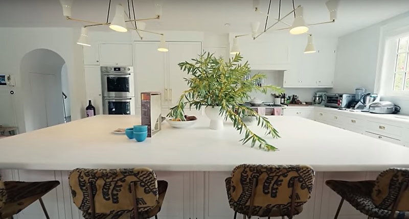 Large white kitchen anchored by a massive marble kitchen island, and featuring gold access in the light fixtures and cabinets, with a plant in the middle of the island.