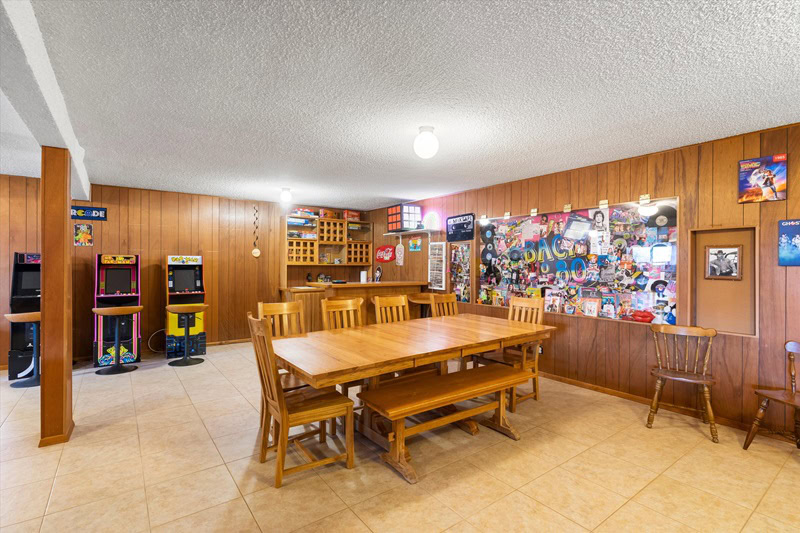 Entertainer's basement with wood paneling, a large table, gaming machines, and a wall with pop culture clippings.