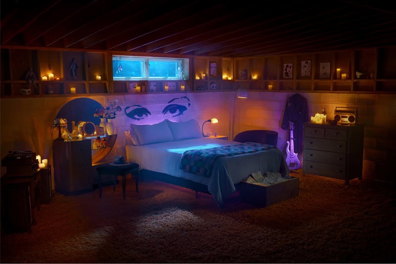 purple-lit bedroom inside Prince's Purple Rain house in Minneapolis, showing a bed with Prince's eyes drawn above it in black ink, with a purple guitar next to the dresser. 