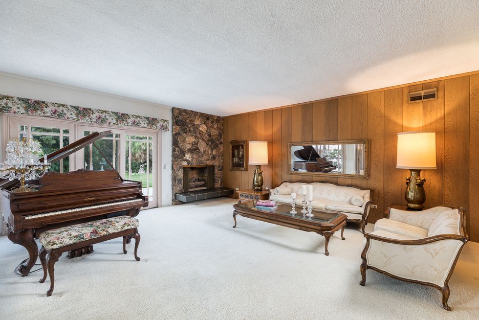Inside the living room of the iconic family home, with a wood-paneled wall, a stone fireplace, elegant seating, and a grand piano
