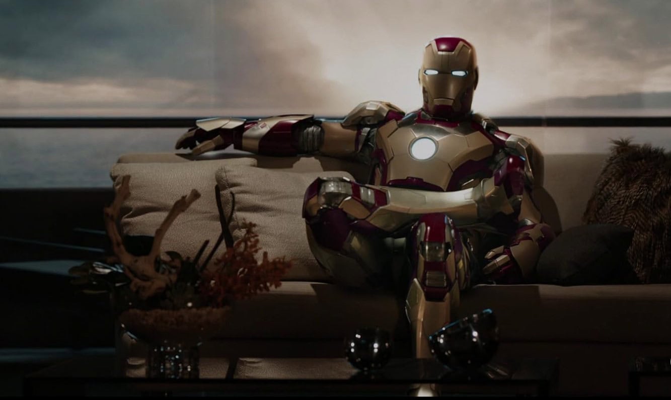 Tony Stark wearing his Iron Man suit and lounging comfortably on the sofa of his house.