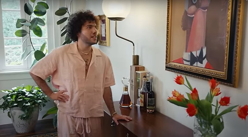 Benny Blanco wearing a light pink outfit looking at a piece of abstract art hung on the walls of his home, with potted plants visible in the background.
