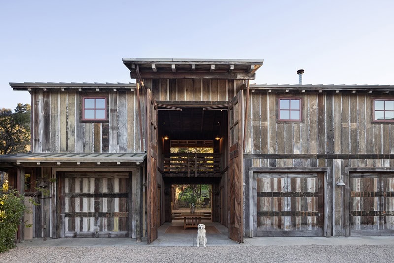a barn made out of reclaimed wood, with its door wide open, showing a table inside. A cream-colored dog stands at the entrance of the barn.