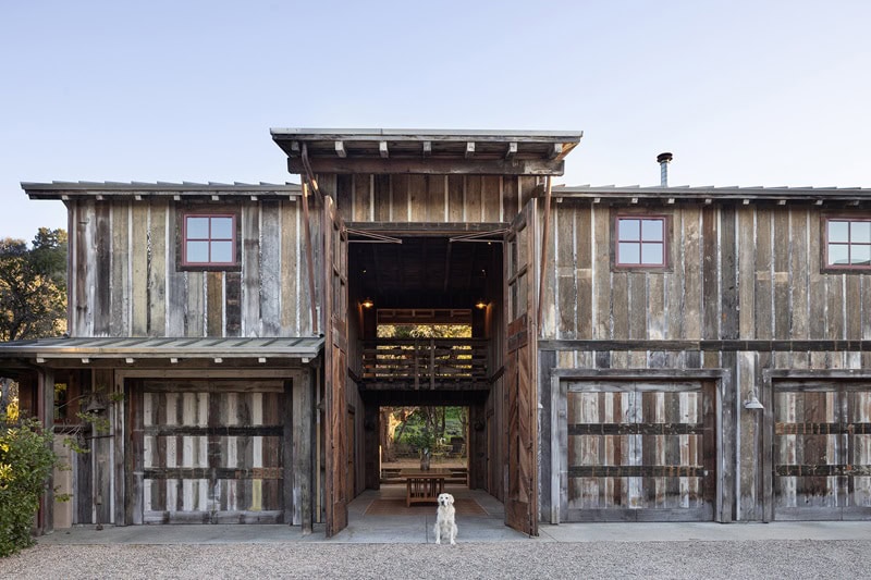 a barn made out of reclaimed wood, with its door wide open, showing a table inside. A cream-colored dog stands at the entrance of the barn. 