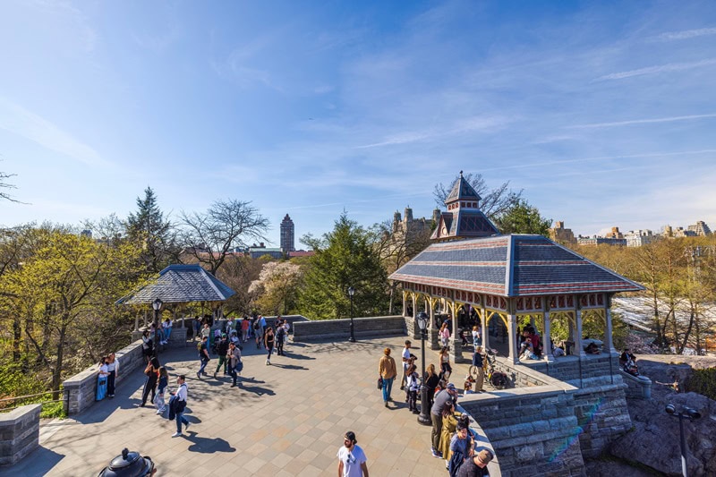 Tourists on the Belvedere Castle terrace in Central Park, New York City, with the crowds admiring the landmarks on a sunny spring day.